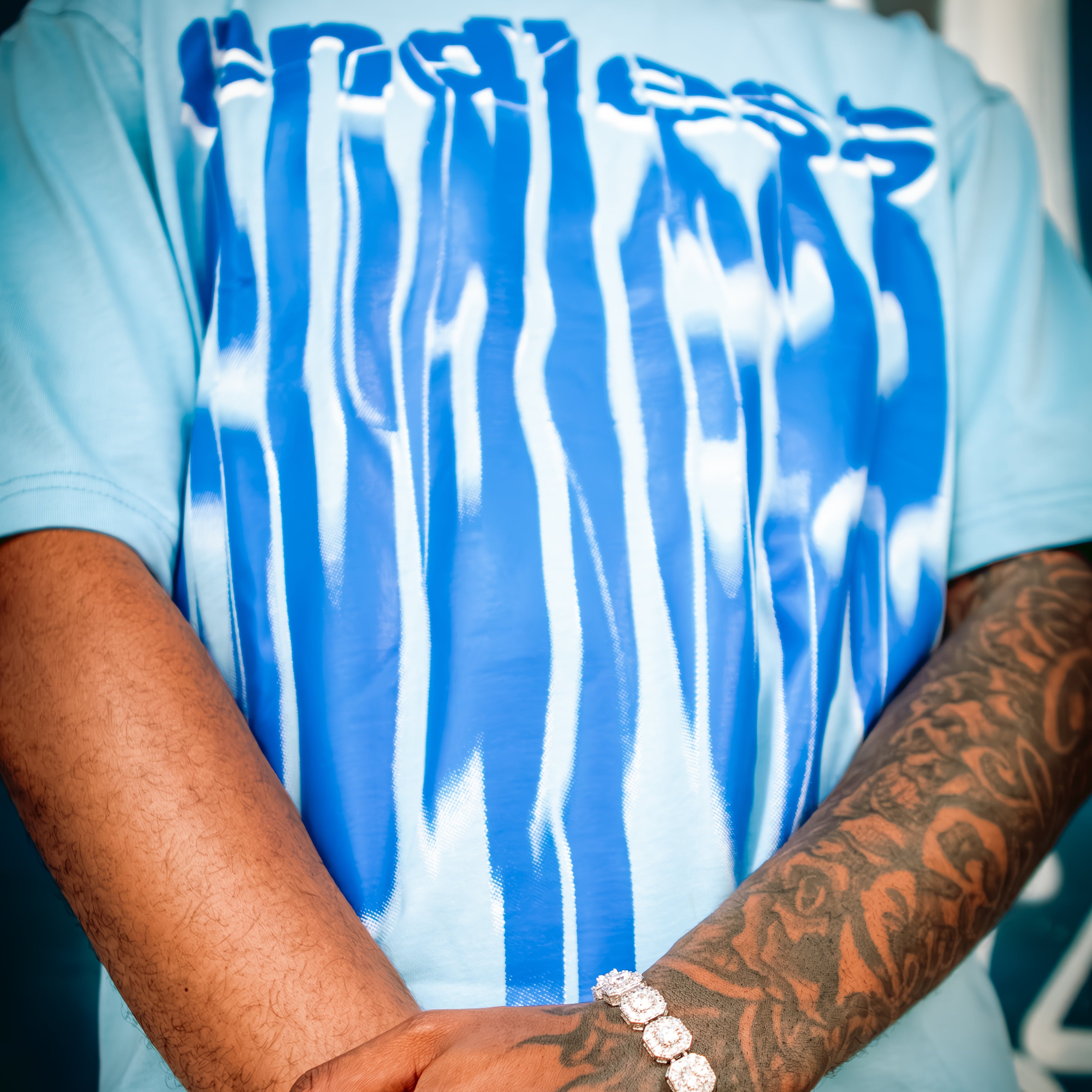 Person wearing a light blue t-shirt with white abstract design, showing tattoos on arms.