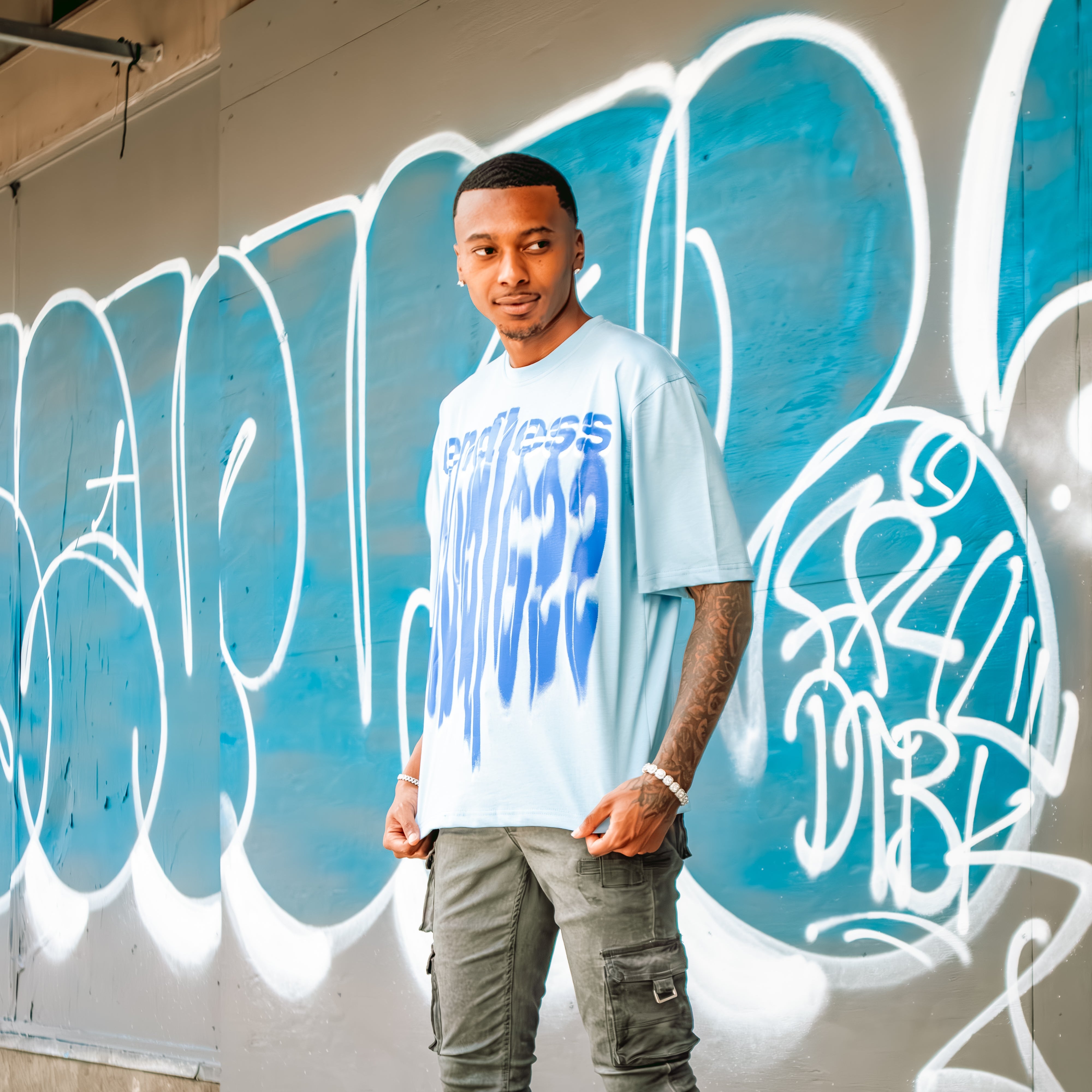 Man wearing a white t-shirt with blue text, gray pants, and blue sneakers standing in front of graffiti.