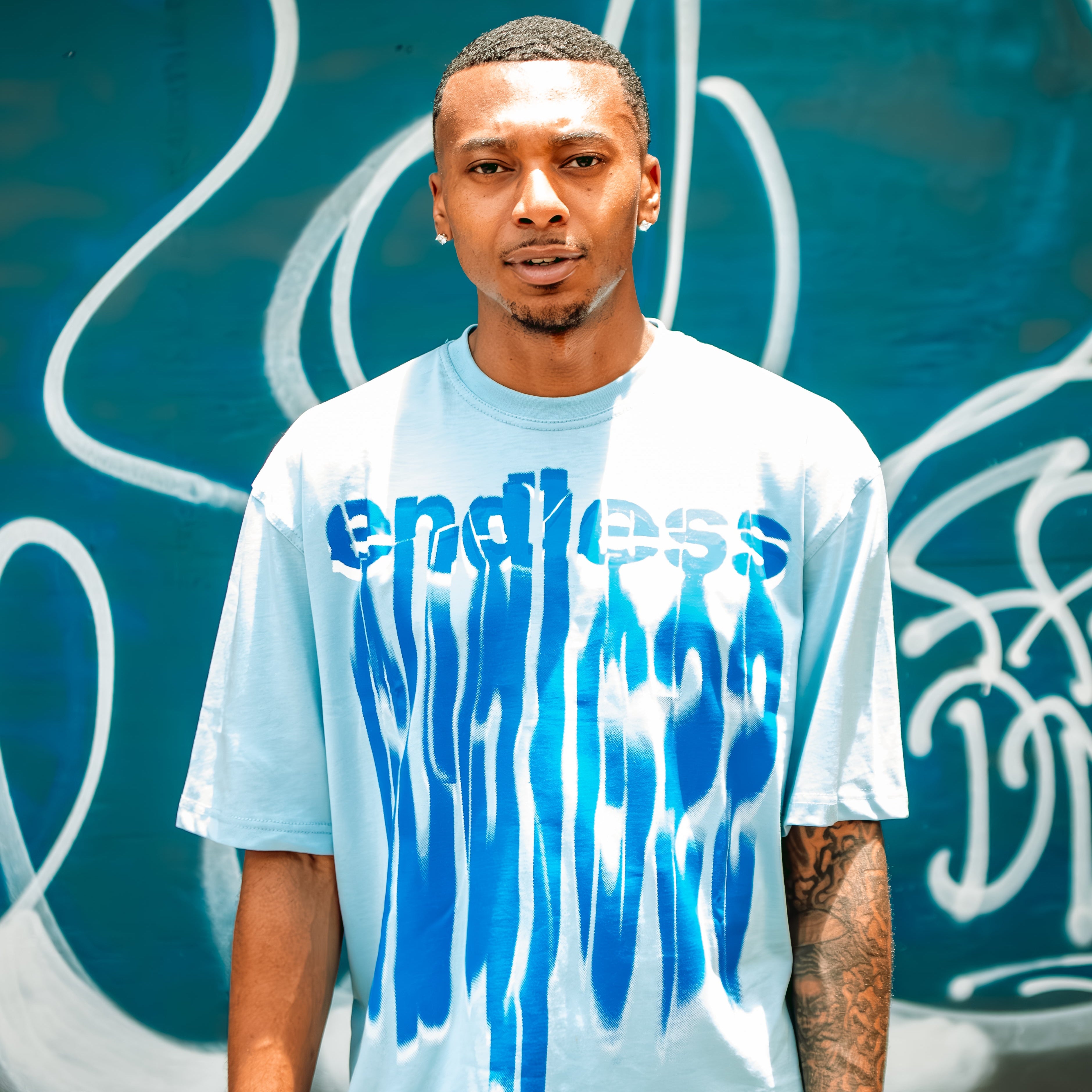 Person wearing a white t-shirt with blue graphics and text, standing against a graffiti-covered wall.